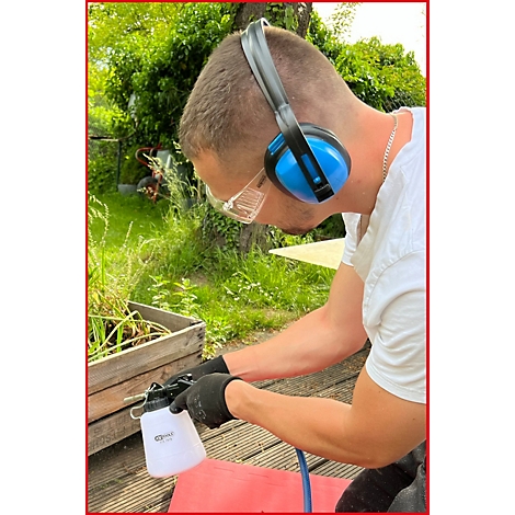 Ein Mann mit Gehörschutz und Schutzbrille hält eine Sprühflasche. Er trägt Handschuhe, im Hintergrund ist ein Garten.